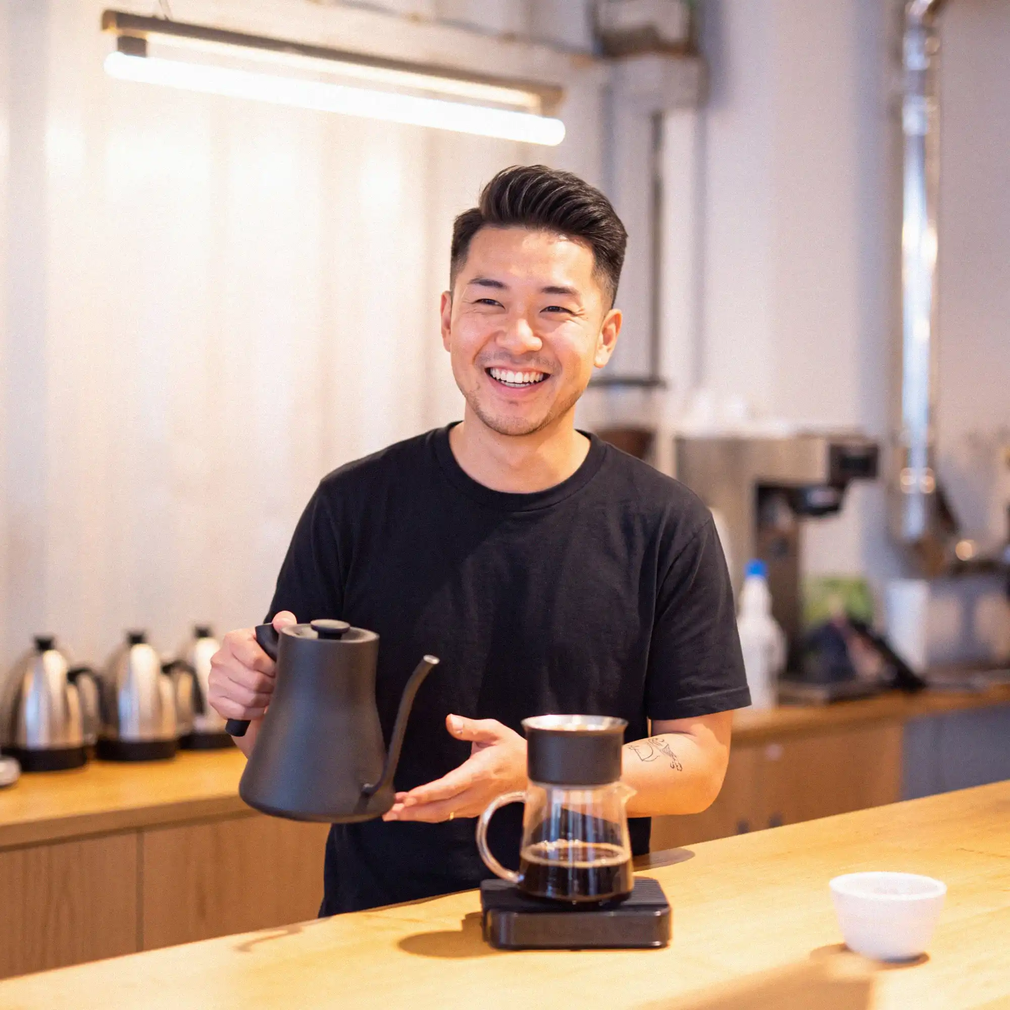 A barista smiling behind the counter of a coffee shop, pouring a filter coffee